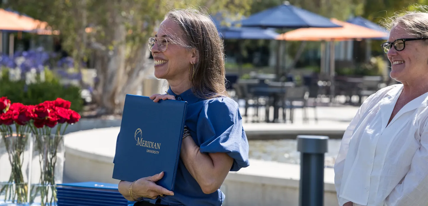 A smiling Meridian graduate holding a Meridian University diploma cover